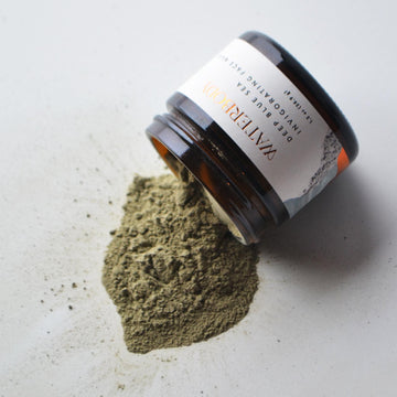A brown glass jar of Waterbody Deep Blue Sea Face Mask is open and spills out over a white surface. The mask is a dry sea green powder